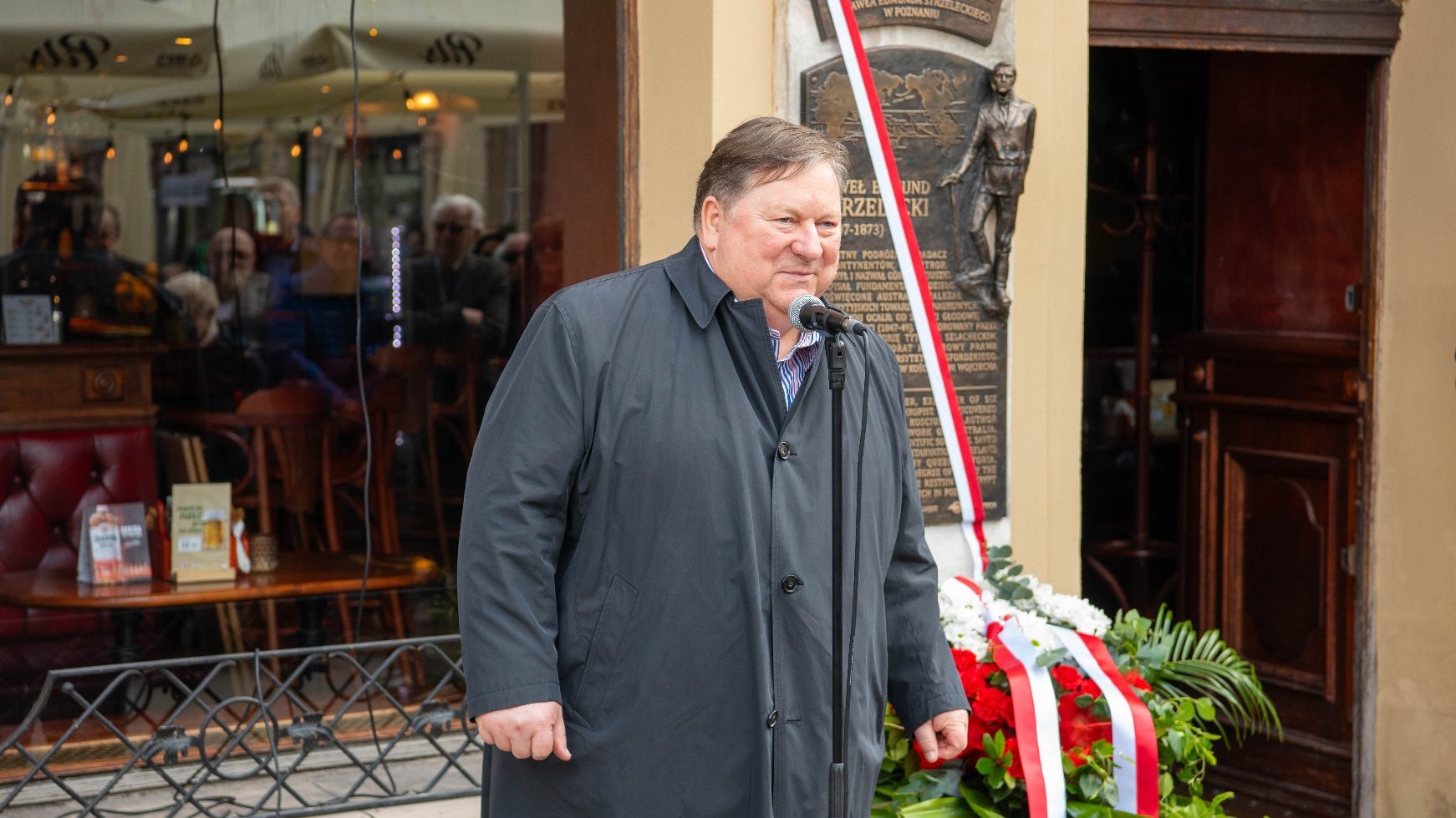 Osoba przemawia do mikrofonu, trzymając kartkę papieru, stoi przed budynkiem przy tablicy pamiątkowej. Tablica zawieszona na ścianie przedstawia postać mężczyzny oraz tekst opisujący jego dokonania. Ozdobiona jest wstęgą w kolorach polskiej flagi (biało-czerwona). U dołu tablicy znajduje się wieniec kwiatowy w kolorach białym i czerwonym, zdobiony wstążką w barwach polskiej flagi.