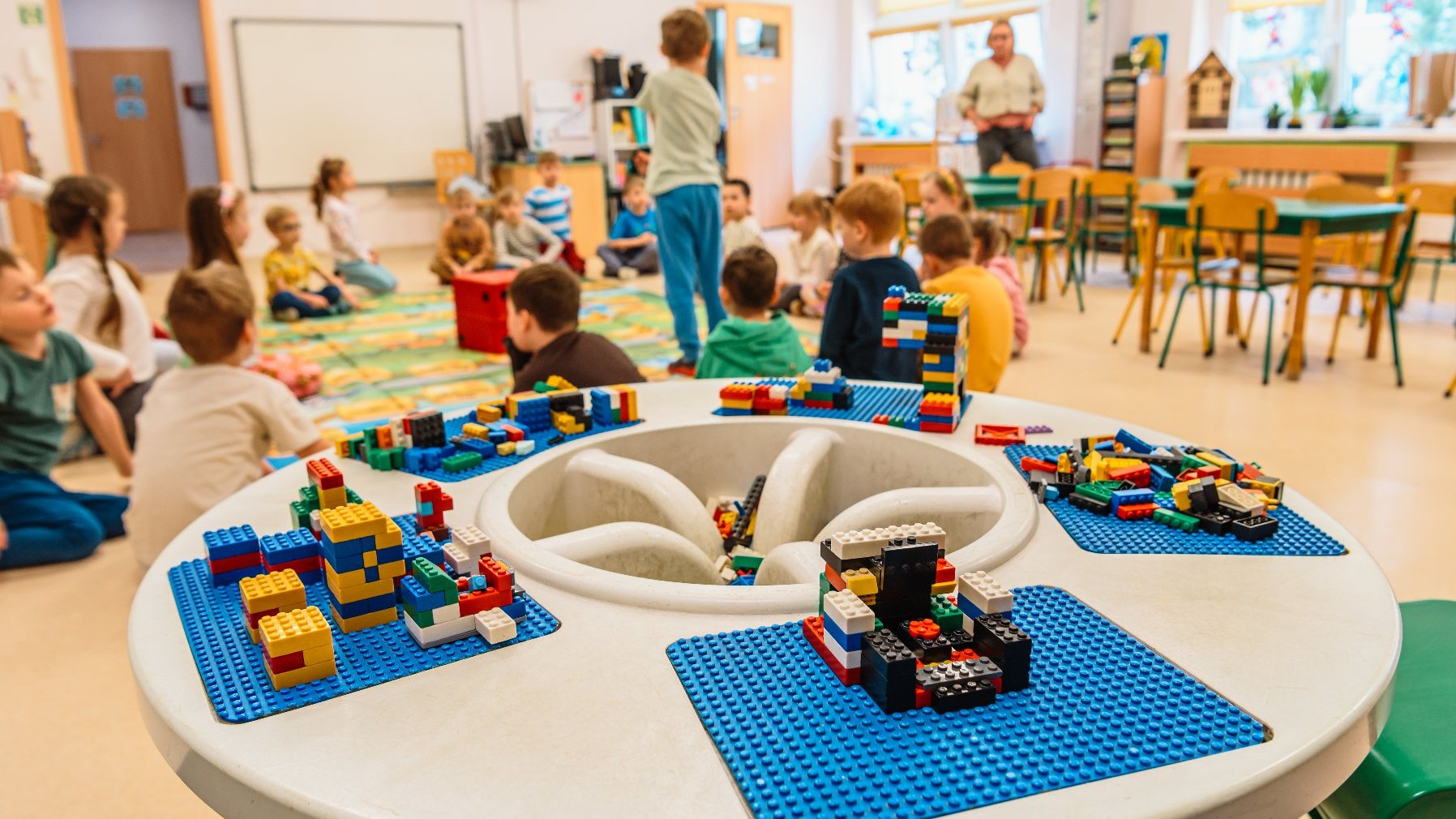 Zdjęcie przedstawia dzieci w sali przedszkolnej siedzące na dywanie. Na pierwszym planie znajduje się stół, na którym leżą klocki lego.