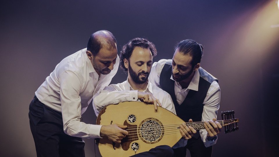 Kolorowe zdjęcie przedstawia trzech mężczyzn, członków zespołu, skupionych wokół jednego instrumentu. Jeden z mężczyzn siedzi, w rękach trzyma lutnię. Dwóch pozostałych jest pochylonych nad instrumentem.