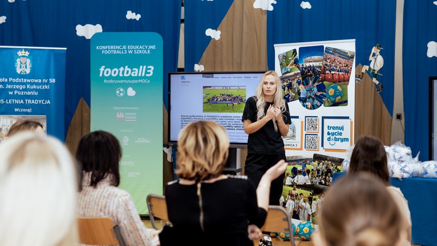 Konferencja edukacyjna "Football3 w szkole 2025" w Poznaniu, fot. Thiem.Photography - prelekcja jednej z prelegentek, która stoi na tle roll-upów reklamujących konferencję oraz ekranu, na którym wyświetlane są materiały. Na pierwszym planie siedzą uczestniczki konferencji