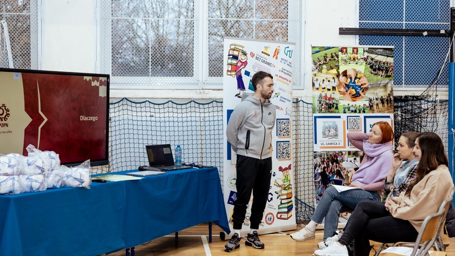 Konferencja edukacyjna "Football3 w szkole 2025" w Poznaniu, fot. Thiem.Photography - prelekcja jednego z prelegentów, który stoi na tle roll-upów i ekranu z prezentacją, zaś prezentacji przysłuchują się trzy siedzące panie