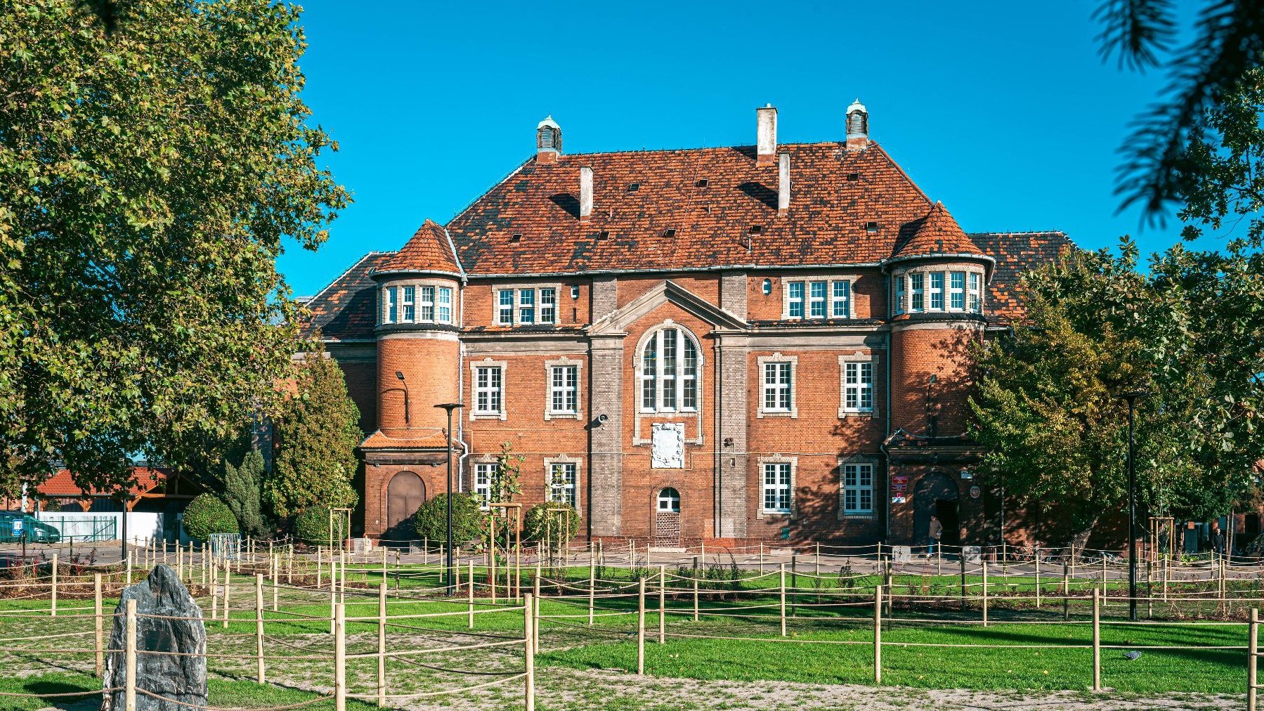 Widok na budynek szkoły. Przed nim zielony trawnik z nowo posadzonymi drzewami.