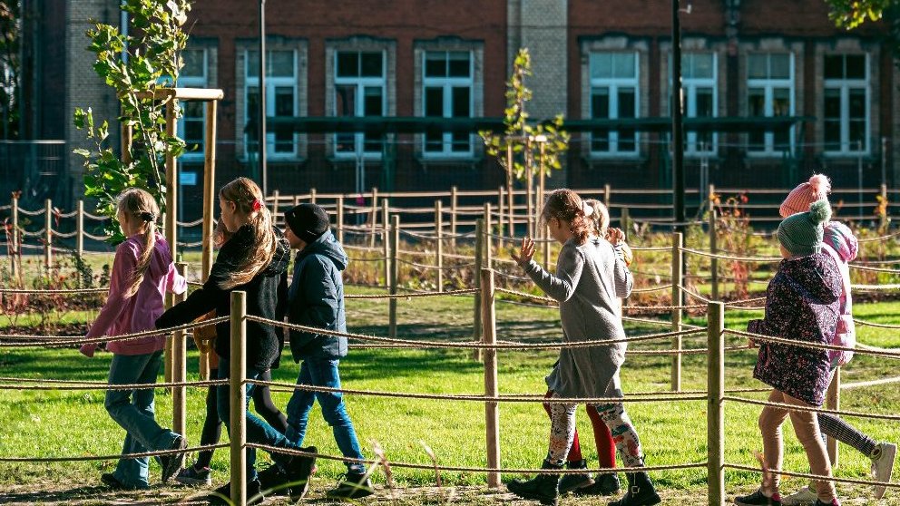 Dzieci na spacerze przechodzące dziedzićcem szkoły.W tel rośliny, zielone drzewa i budynek szkolny.