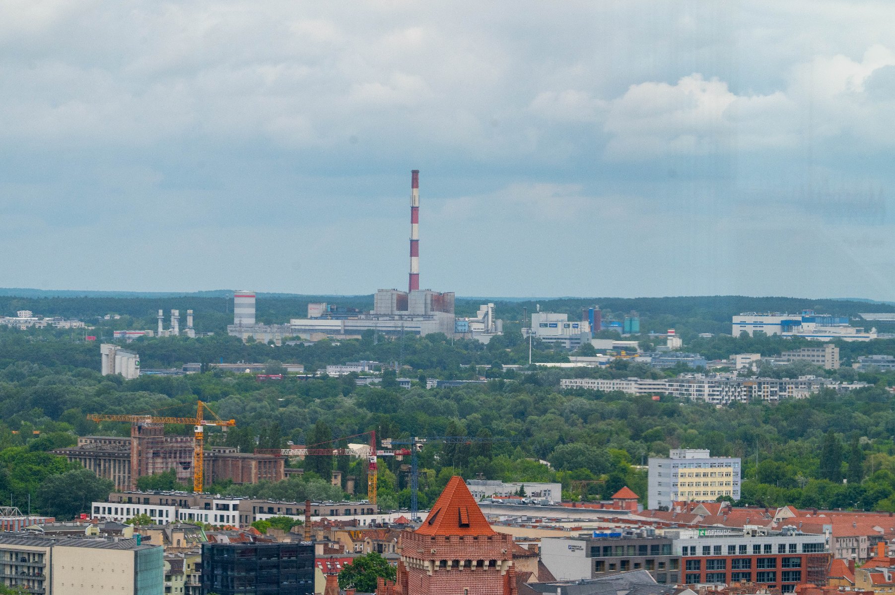 Celem konsultacji jest zebranie opinii, uwag, potrzeb oraz obaw poznaniaków w zakresie lokalnej polityki energetycznej Panorama Poznania. Na pierwszym planie dachy budynków i szczyt wieży kościelnej. W tle widoczny jest las oraz zabudowania miejskie, w tym wysoki, biało-czerwony komin elektrociepłowni.
