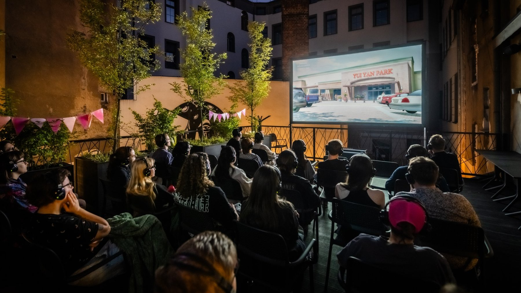 Silent Cinema rusza już 1 sierpnia / fot. Piotr Bedliński Wieczorny pokaz kina plenerowego. Widzowie siedzą na krzesłach, oglądając film wyświetlany na dużym ekranie. Niektórzy mają na sobie słuchawki. Wokół widoczne są drzewa, lampki i dekoracje w postaci chorągiewek.