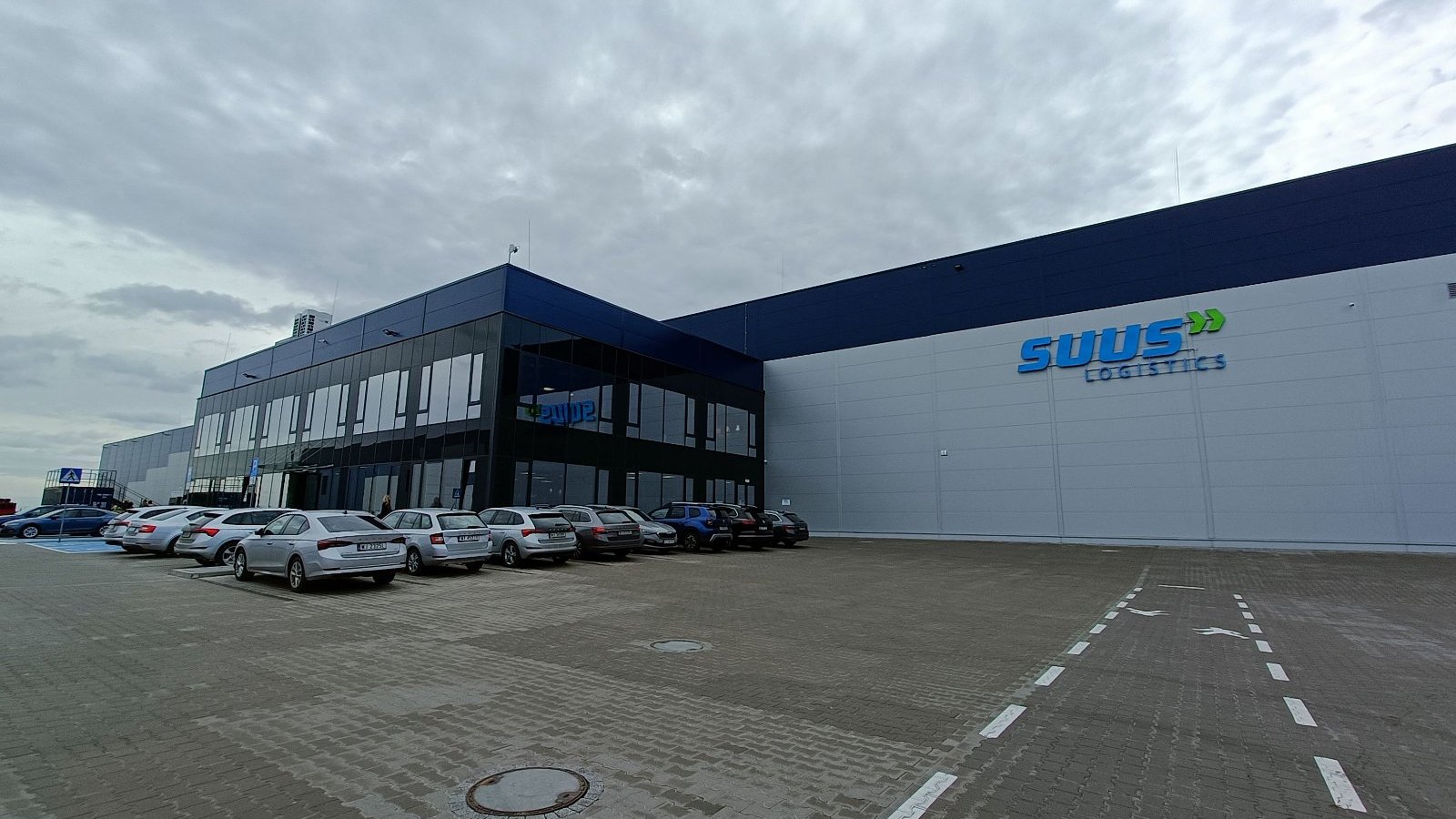 The photo shows the warehouse building of SUUS Logistics. There is a glass entrance in front of the warehouse. Passenger cars are parked in the car park.