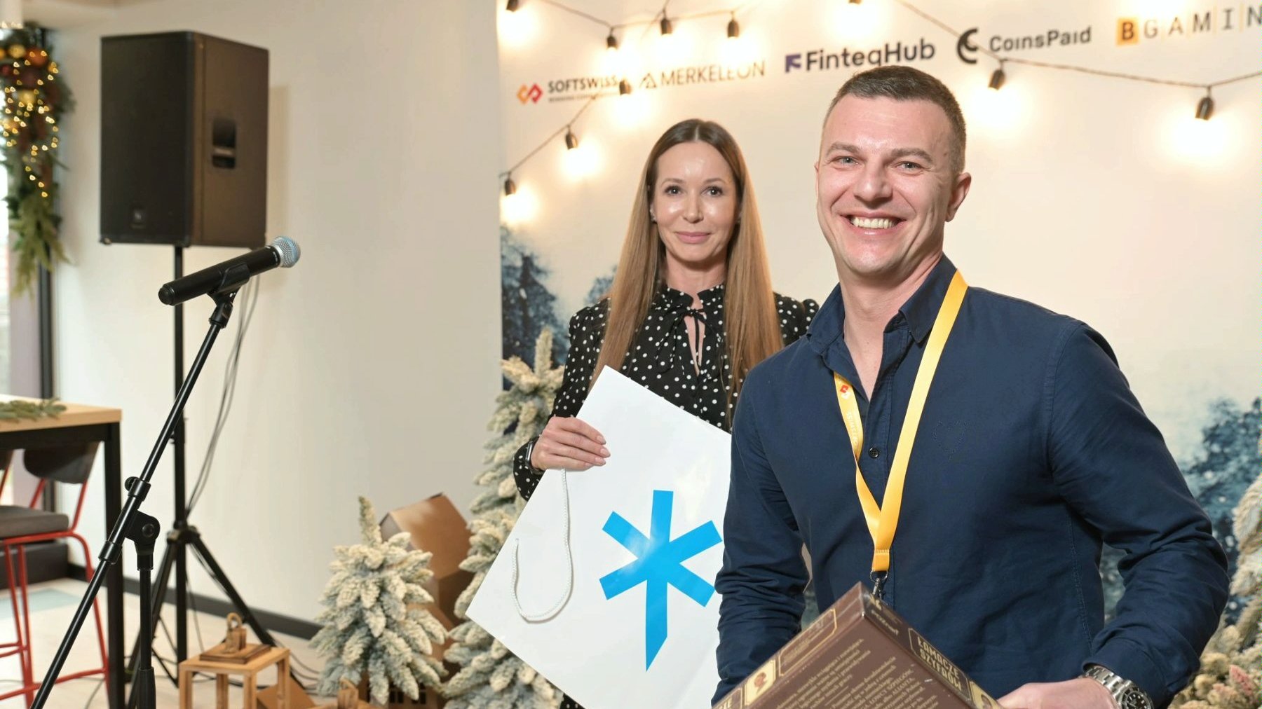 The photo shows Ivan Montik, founder of SOFTSWISS. The man is smiling, holding the board game Code Breakers. Behind him is the director of the Investor Relations Department, Katja Lozina. In the background is a festive wall with company logos.