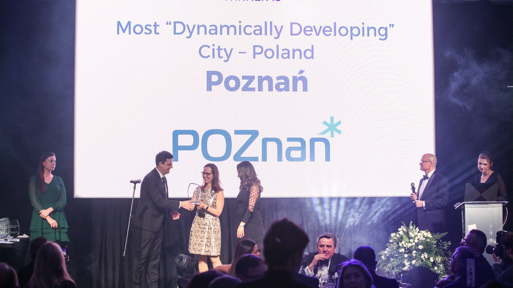 The image is a photograph from the gala. In the background, a large screen shows the caption: "Most Dynamically Developing City - Poland. Poznań'. In front of the screen stand the gala hosts. They are presenting the award to the Investor Relations Department.