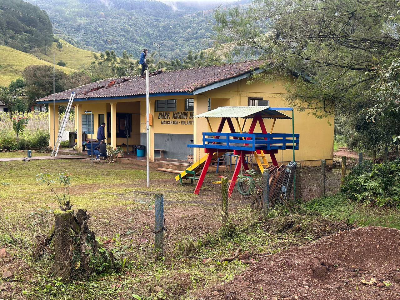 Yellow, low school building in the background of the mountain range, to the right of the picture, next to the school a colourful playground (blue-red-yellow) with a slide and swings - grafika artykułu