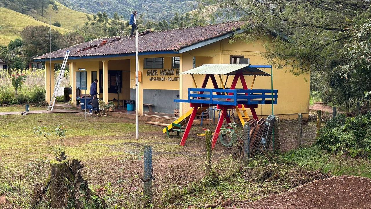 Yellow, low school building in the background of the mountain range, to the right of the picture, next to the school a colourful playground (blue-red-yellow) with a slide and swings