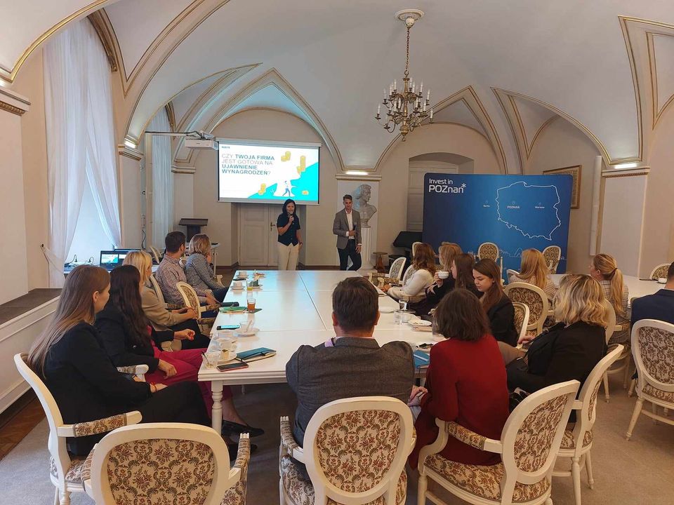 In the background is the presentation "Is your company ready for salary disclosure, on the right is the dark blue Invest in Poznan wall with white lettering and the outline of the map of Poland, in front you can see people listening in - grafika artykułu