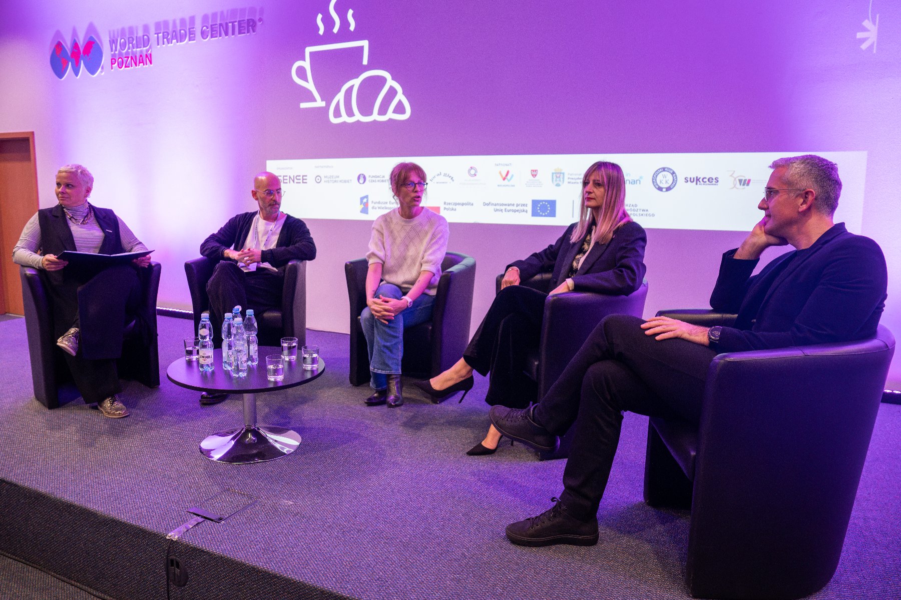 The photo shows 5 people sitting at a table with water: from left, a woman with gray hair holding a notebook on her lap, probably a man with glasses with stubble on his face and no hair on his head, a woman with her hands braided on her legs, a woman resting her hands on the back of a chair, a man with glasses, supporting his head with his hand, with a short haircut, an inscription can be seen in the upper left corner: World Trade Center Poznań - grafika artykułu