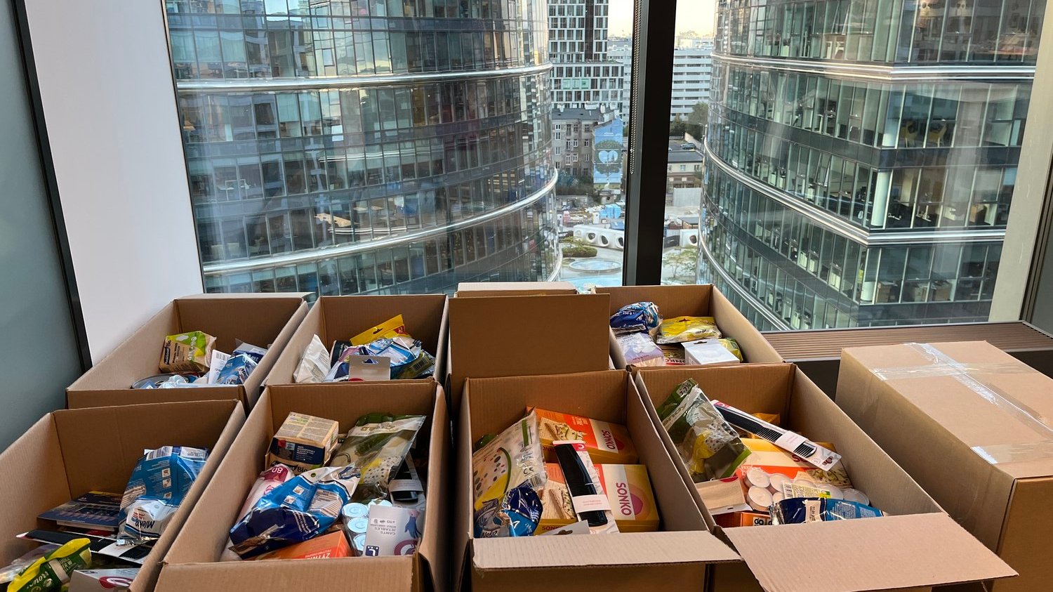 Cartons of food and hygiene products for flood victims. The stacked cardboard boxes lie under the window, with office buildings visible in the background.