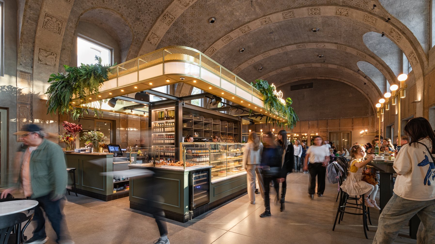 The interior of the Petit Paris café in Betonhaus. People are sitting at tables, eating their meals. Sweet baked goods can be seen behind the counter.