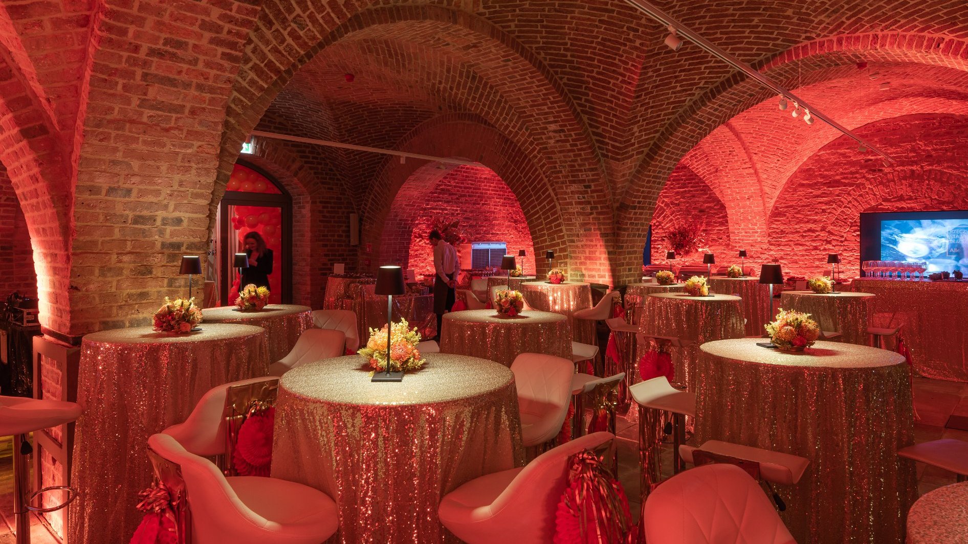 The great hall in the basement of the former brewery. Festive tables are decorated with flowers. A projector can be seen in the distance.