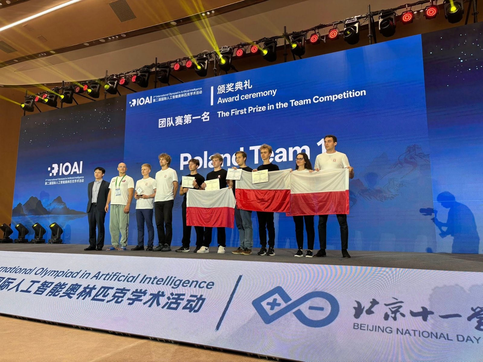 Polish team at the International Olympiad in Artificial Intelligence in Beijing. Photo: International Olympiad in Artificial Intelligence The Poland team poses on stage with flags. In the background, the words: "The First Prize in the Team Competition".