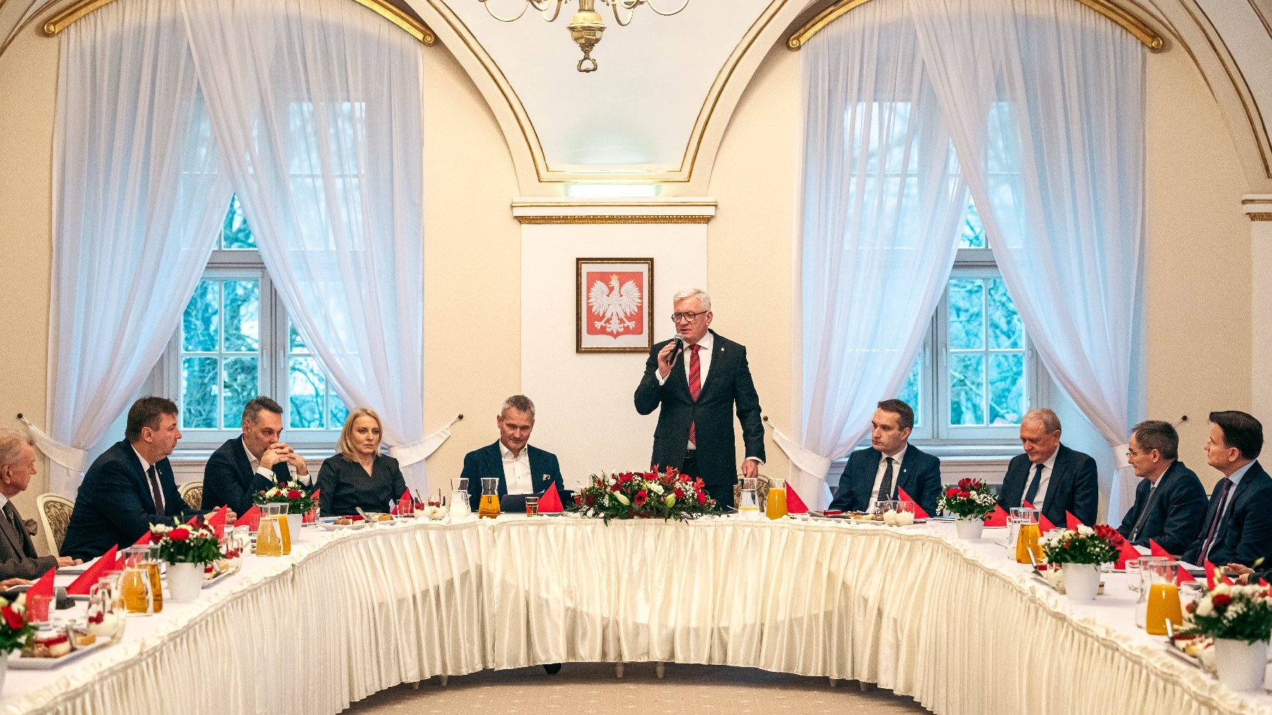 Formalne spotkanie w eleganckim pomieszczeniu z białymi zasłonami, żyrondami i dużą lampą wiszącą na suficie. Na środku znajduje się stoł z białym fartuchem, czerwonymi ręcznikami, kwiatami i dzbankami z napojami. Wokół stołu siedzą osoby, a jedna osoba stoi i wydaje się prowadzić prezentację lub mówić do grupy. Na ścianie znajduje się ujęte w ramę godło Polski - biały orzeł na czerwonym tle.