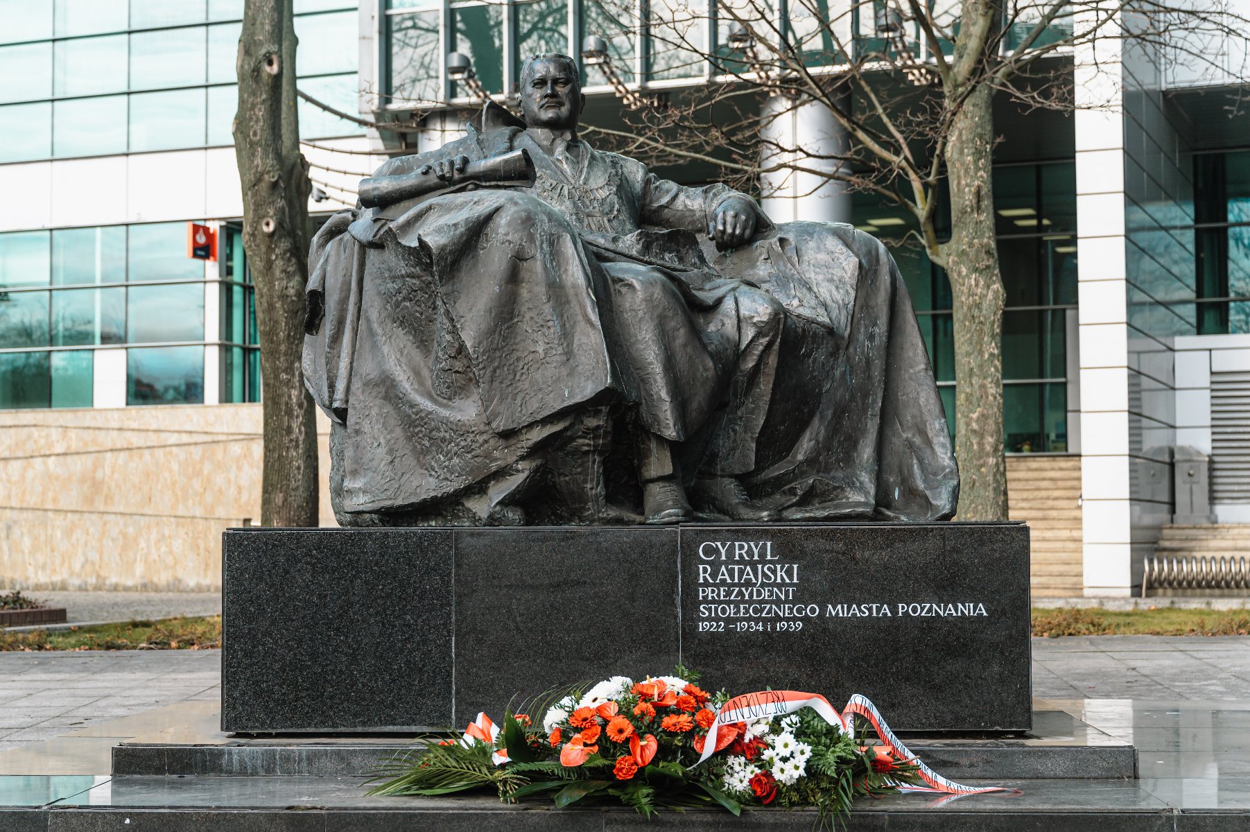 Cyryl Ratajski siedzący na pomniku z brązu na czarnym cokole. Przed cokołem ułożone są kwiaty w kolorach białym, czerwonym i zielonym oraz wstążka. Na cokole widnieje napis: "CYRYL RATAJSKI PREZYDENT STOŁECZNEGO MIASTA POZNANIA 1922 - 1934 I 1939". - grafika artykułu