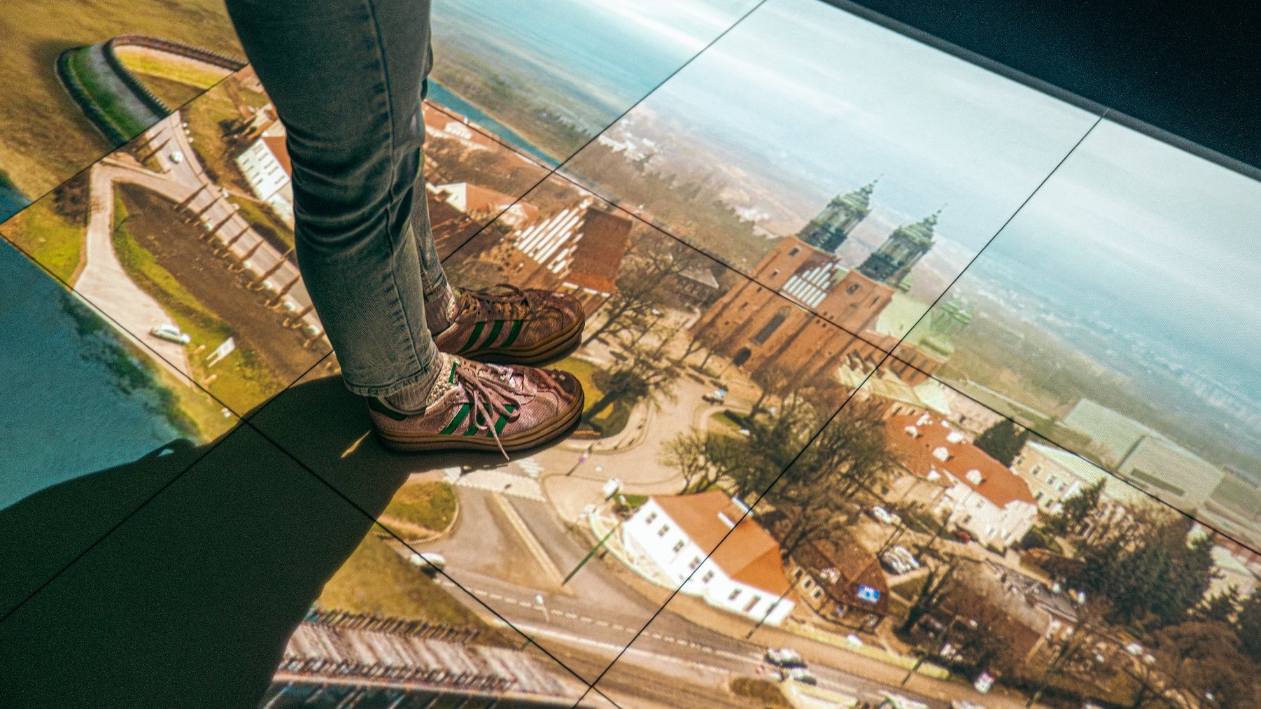 Zdjęcie przedstawia fotografię Ostrowa Tumskiego, m.in. katedry poznańskiej na podłodze oraz nogi osoby, kóra na nim stoi.