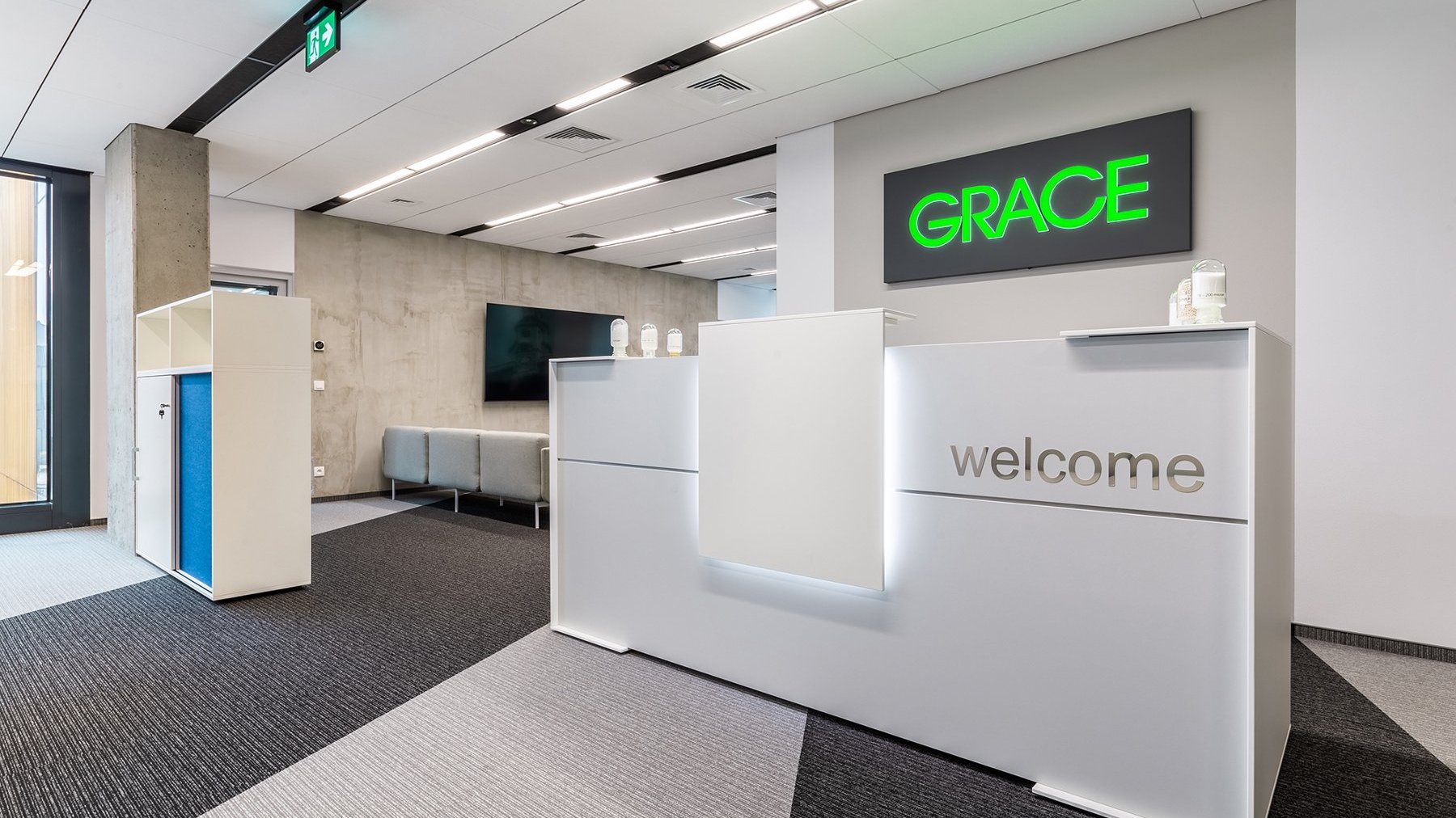 Zdjęcie przedstawia białą ścianę, widnieje na niej logo firmy Grace. Przed nim znajduje się biała recepcja z napisem "Welcome".