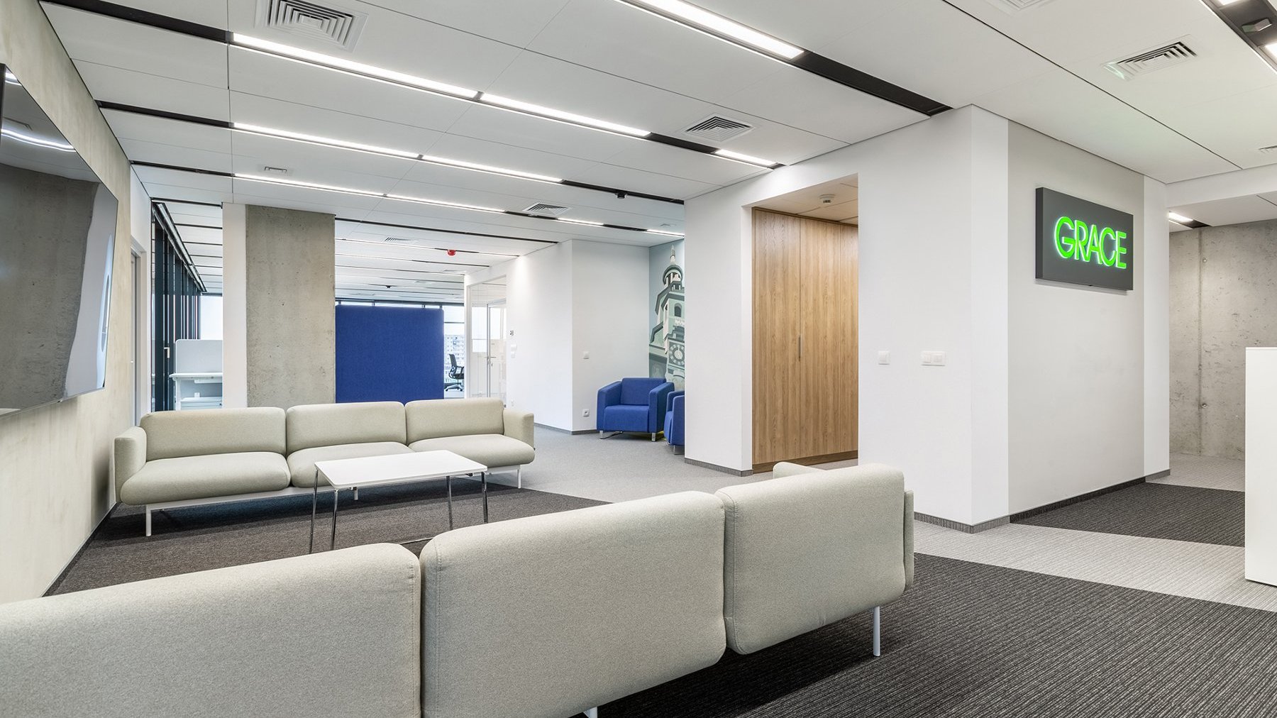 Zdjęcie przedstawia nowoczesną przestrzeń biurową. Po prawej stronie na ścianie wisi neonowe logo firmy Grace. Po lewej naprzeciwko siebie stoją dwie materiałowe kanapy. W tle widać pomieszczenia biurowe.