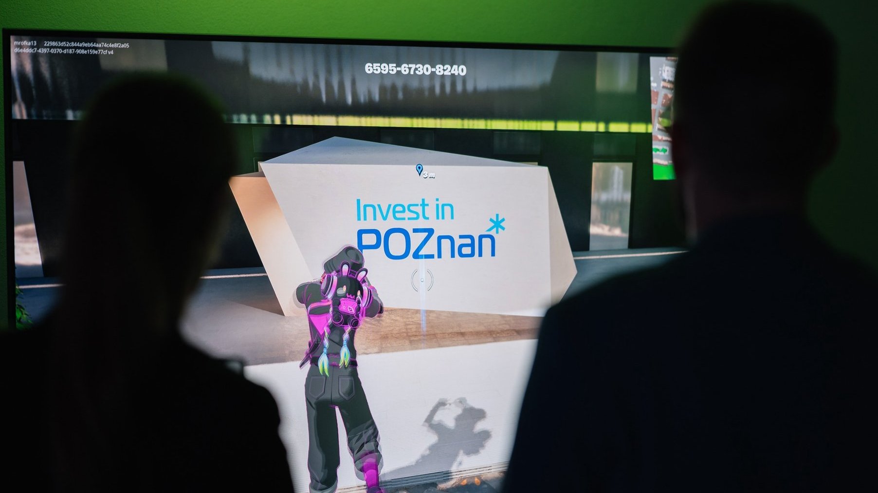 Zdjęcie przedstawia dwie osoby stojące plecami do kamery. Obydwie zwrócone są w kierunku ekranu, na którym włączona jest gra Fortnite. Postać w grze patrzy na monument z logotypem Invest in Poznań.