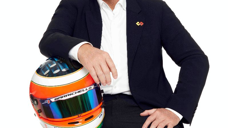 Rubens Barrichello na białym tle ubrany w garnitur. Uśmiechnięty, trzyma pod pachą kask motorowy.