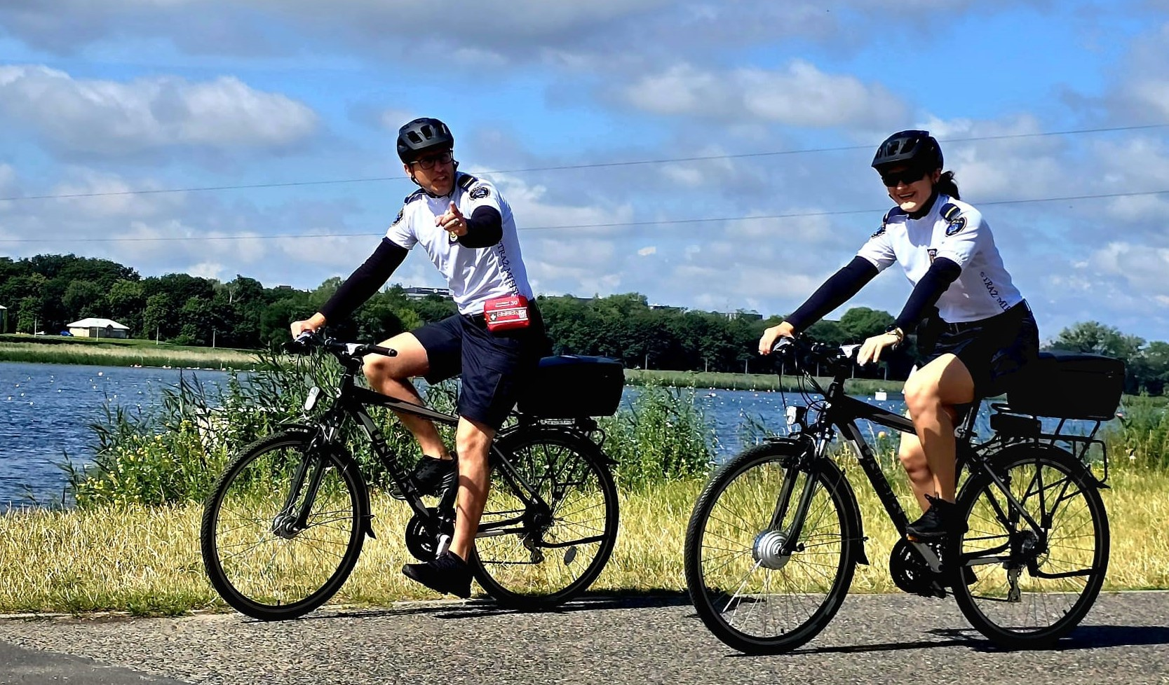 Patrole rowerowe kierowane są przede wszystkim do parków i w okolice Wartostrady Dwoje rowerzystów, prawdopodobnie policjantów lub strażników miejskich, w czarnych kaskach i białych koszulkach z długimi rękawami, jedzie na rowerach po ścieżce rowerowej. Obok nich znajduje się jezioro lub rzeka, a w tle widać zieleń i błękitne niebo z chmurami. Mężczyzna po lewej stronie wskazuje palcem w kierunku widza, kobieta za nim usmiecha się..