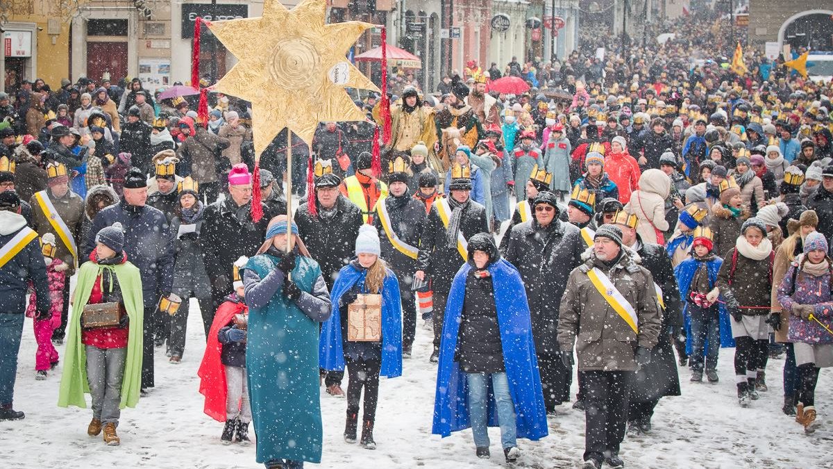 Zdjęcie przedstawia tłum ludzi idących przez Stary Rynek. Osoby w pierwszym rzędzie mają przebrania i niosą dużą złotą gwiazdę. Pada śnieg.