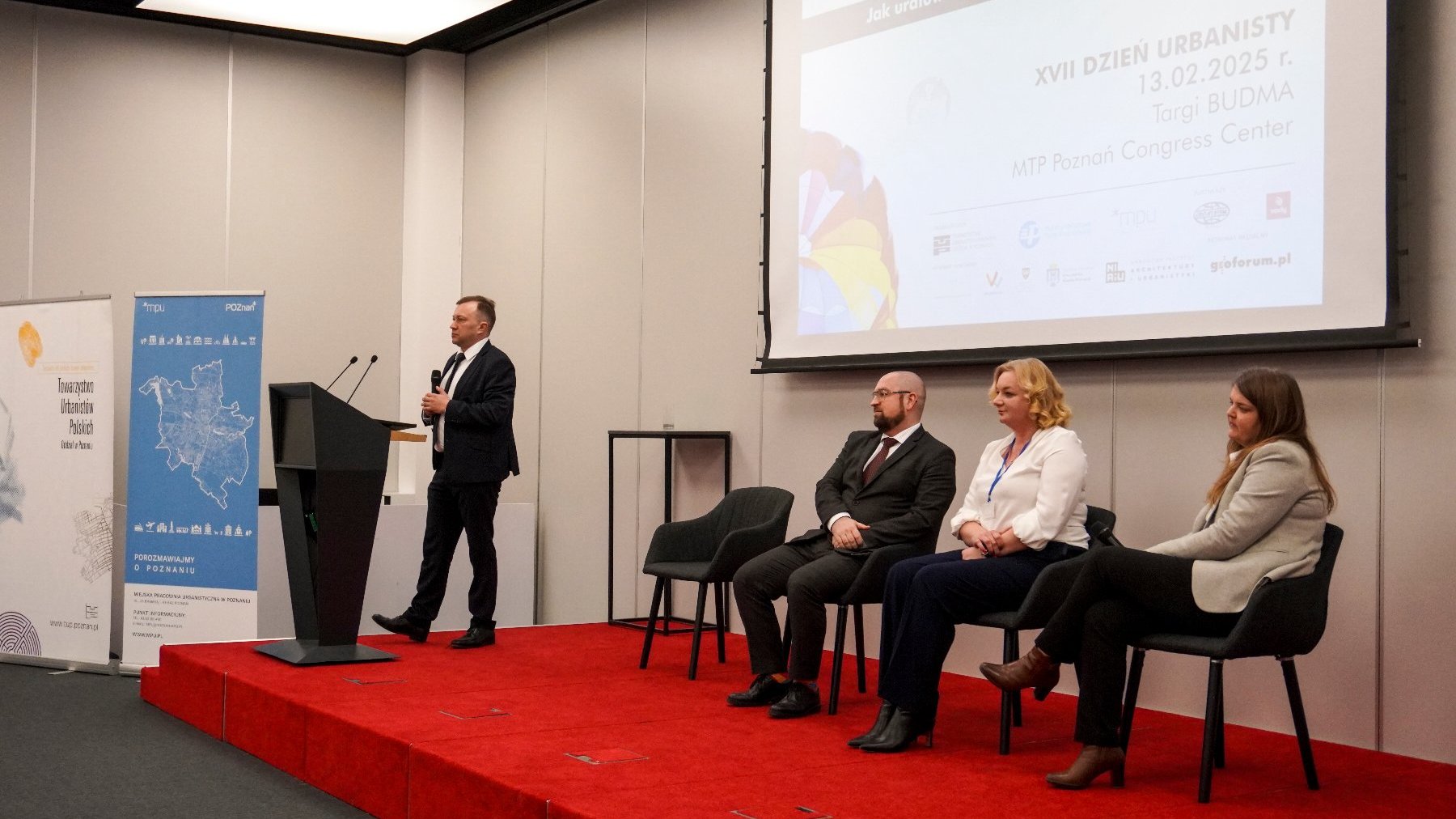 Konferencja lub seminarium z udziałem czterech osób na scenie. Jedna osoba stoi przy mównicy i mówi do mikrofonu, podczas gdy trzy pozostałe siedzą na krzesłach na scenie. Za nimi znajduje się duży ekran wyświetlający informacje o wydarzeniu.