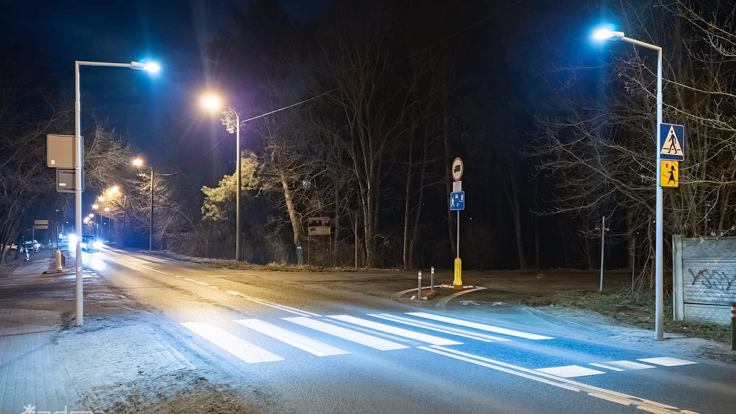 Nocny widok ulicy z oświetlonym latarniami przejściem dla pieszych, wyraźnie widoczna zebra.