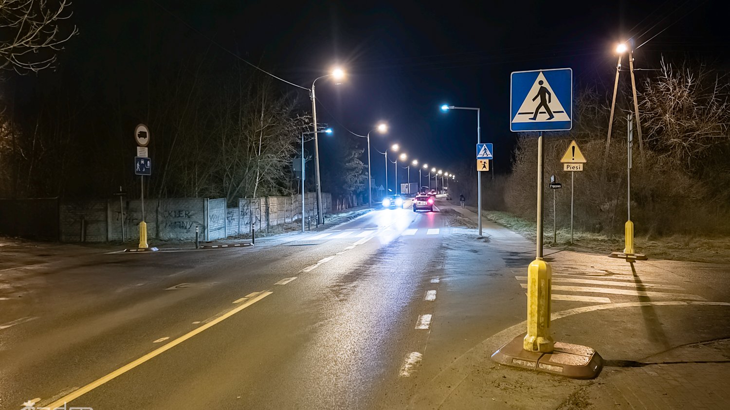 Nocny widok ulicy, na pierwszym planie oświetlone latarniami przejście dla pieszych i znak ostrzegawczy.