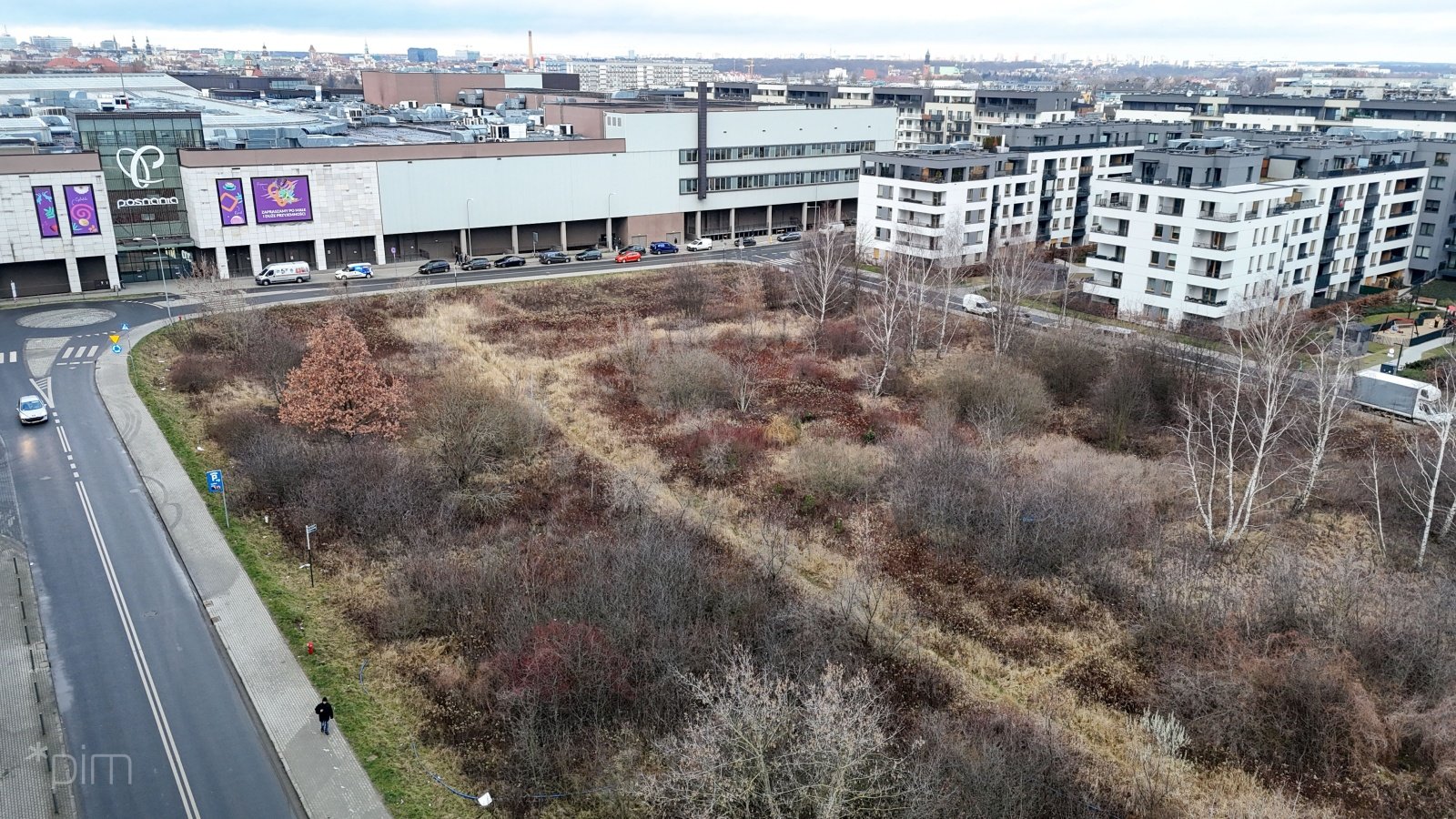 Niezagospodarowana działka porośnięta dziką roślinnością, z przylegającymi do niej jezdniami. Obok intensywana zabudowa nowymi kilkupiętrowymi blokami mieszkaniowymi i centrum handlowe.