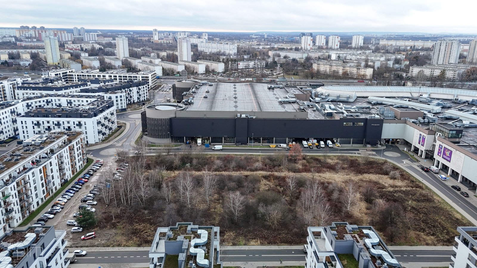 Niezagospodarowana działka pokryta dziką roślinnością. Wokoło ulice miasta, miejsca postojowe oraz gęsta zabudowa mieszkaniowa i centrum handlowe.