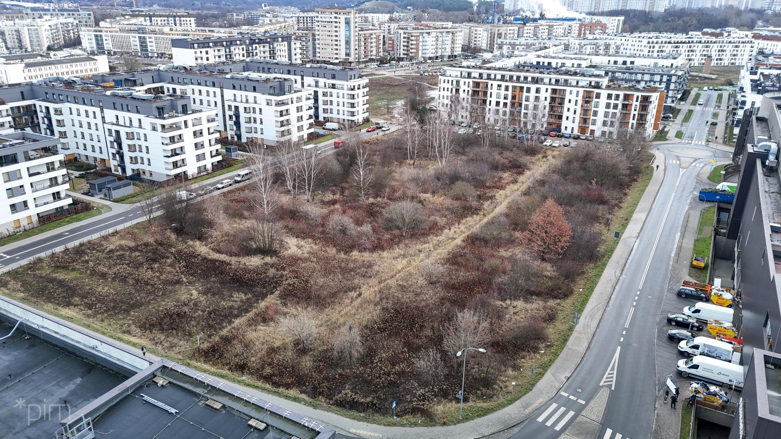 Niezagospodarowana działka porośnięta dziką roślinnością, z przylegającymi do niej z każdej strony jezdniami. Wokoło intensywana zabudowa nowymi kilkupiętrowymi blokami mieszkaniowymi. - grafika artykułu