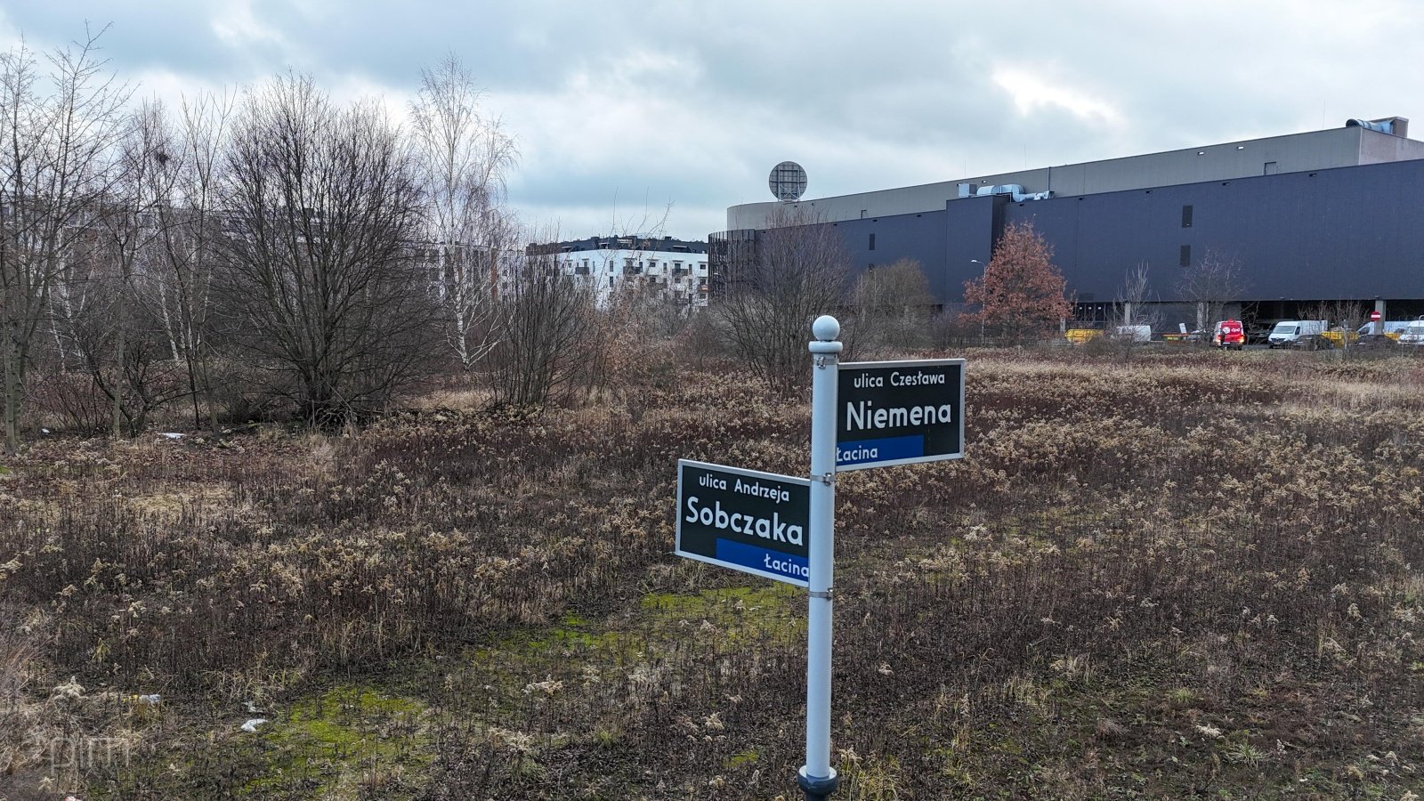 Narożnik niezagospodarowanej działki z dziką roslinnością i słupek z nazwami ulic "Sobczaka" i "Niemena". Obok budynki centrum handlowego.