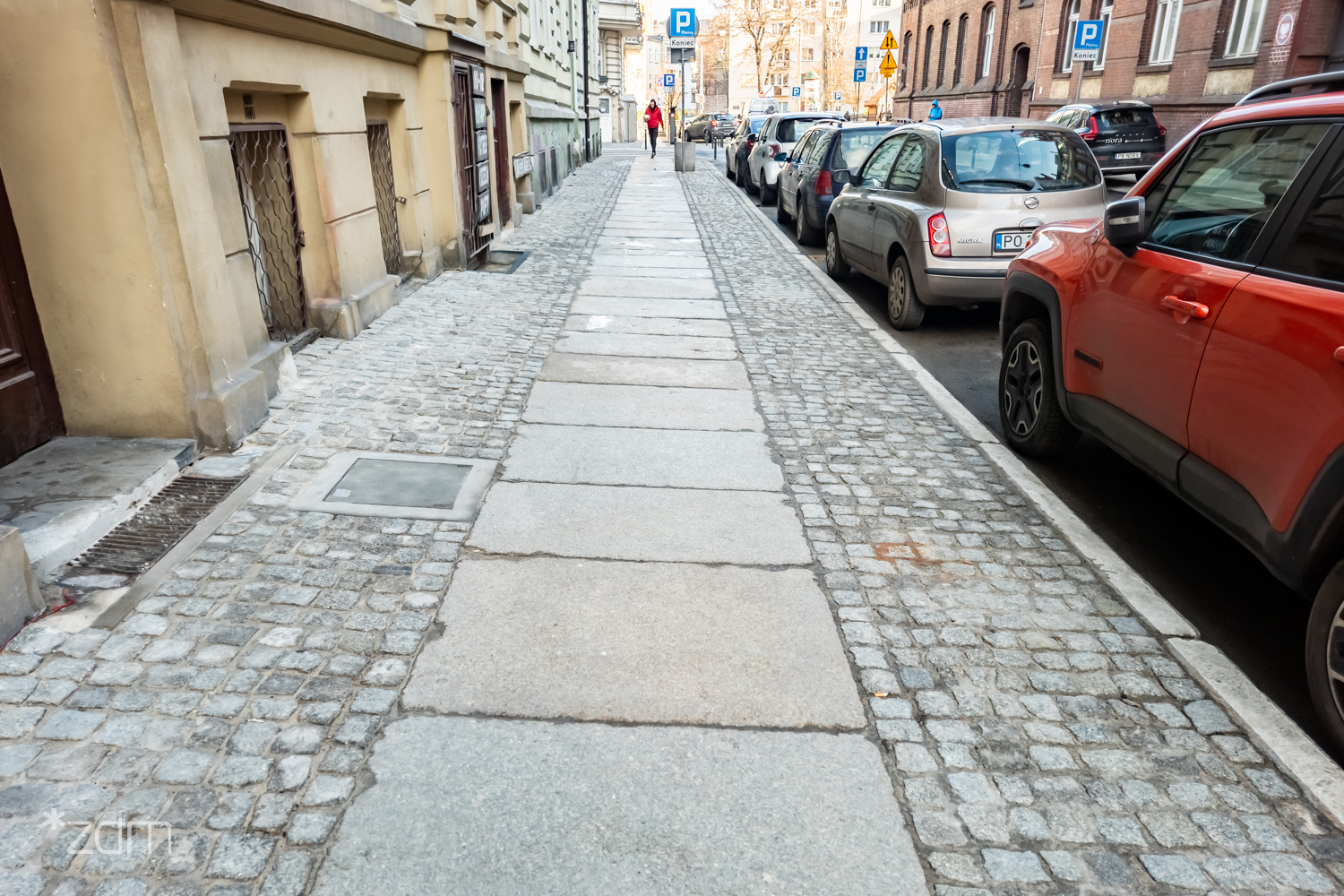 Widok na wyremontowany chodnik z kostki i dużych płyt, obok jezdnia z samochodami i ciąg budynków. - grafika artykułu