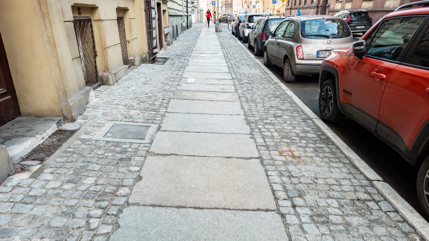 Widok na wyremontowany chodnik z kostki i dużych płyt, obok jezdnia z samochodami i ciąg budynków.