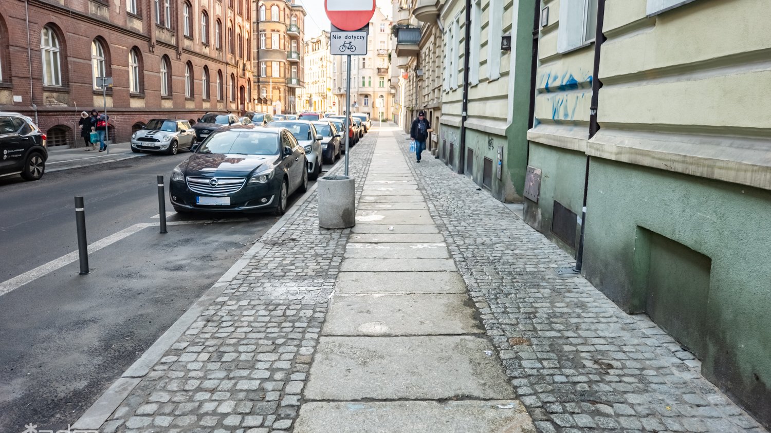 Widok na wyremontowany chodnik z kostki i dużych płyt, obok jezdnia z samochodami i ciąg budynków. Na skraju chodnika znak pionowy i kosz na śmieci.