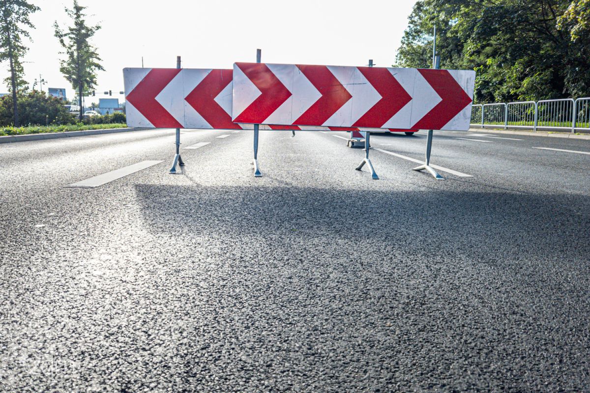 Bariery drogowe w ostrzegawczych biało-czerwonych barwach na środkowym pasie jezdni z nową nawierzchnią asfaltową zabezpieczają prace na dalszym odcinku ulicy i wskazują kierunek jazdy. - grafika artykułu