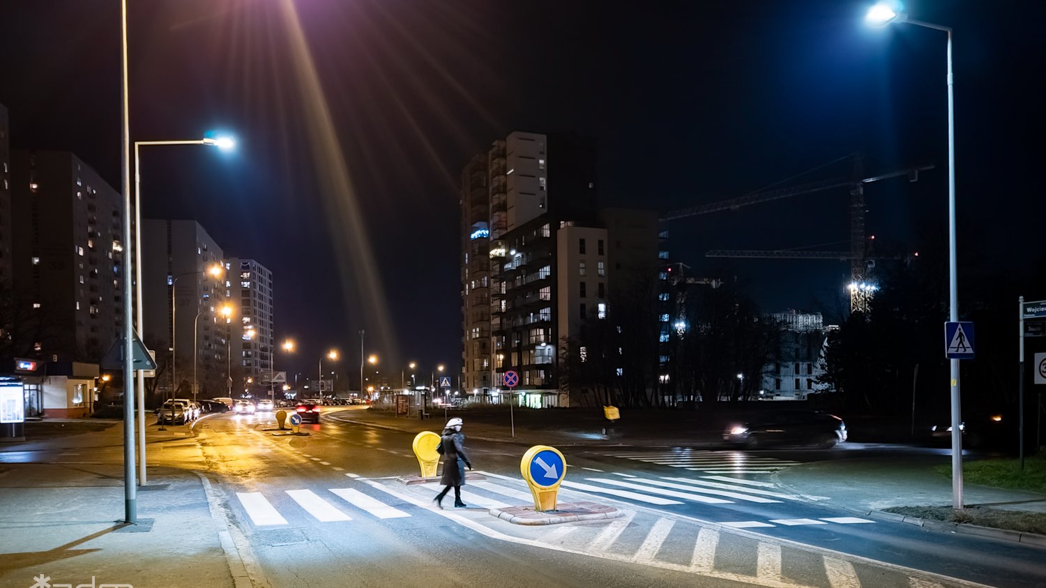 Nocna ulica miasta. Przez oświetlone z obu stron latarniami i oznakowane przejście dla pieszych przechodzi kobieta. Na środku przejścia z wyraźnie widoczną zebrą wygrodzony słupkami azyl.