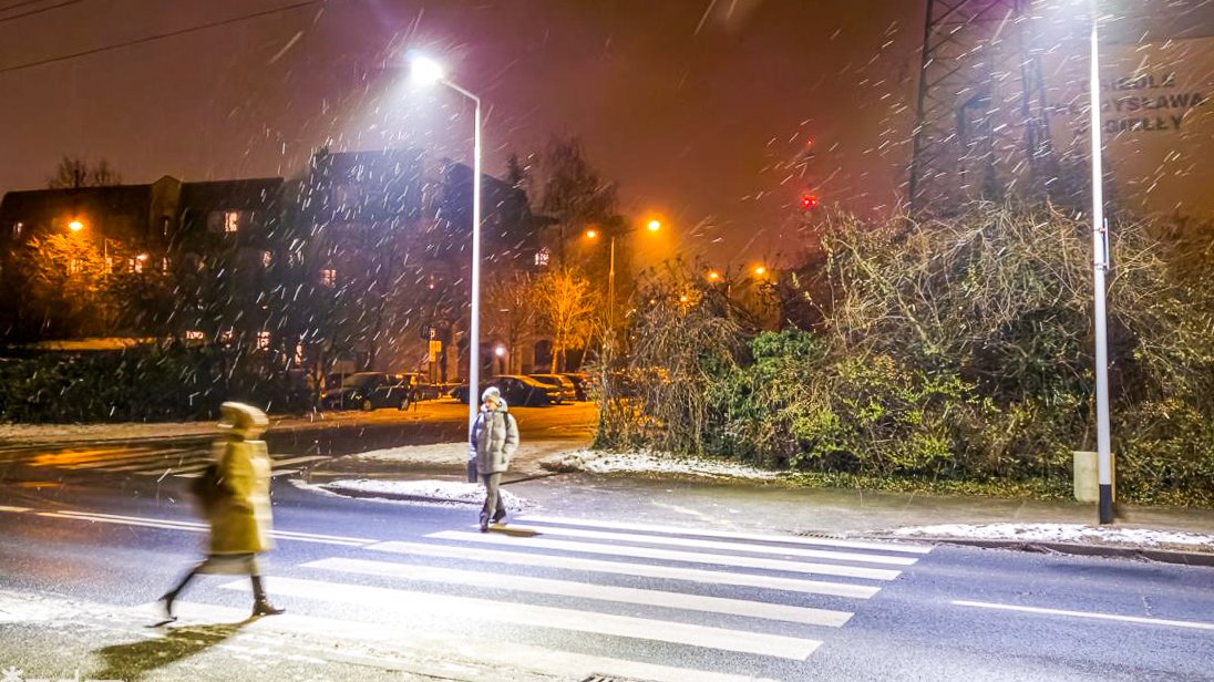 Dwie osoby przechodzą przez doświetlone wieczorem latarniami przejście dla pieszych. Mimo padającego sniegu wyraźnie widoczna zebra i przechodzące przez skrzyżowanie postaci. W tle zieleń przyuliczna i budynki.