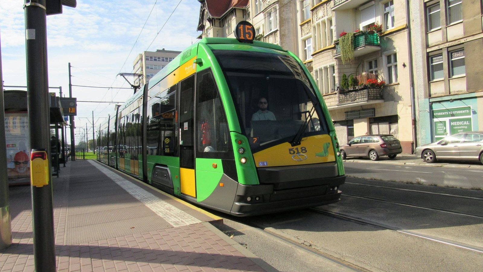 Zielony tramwaj nr 15 na przystanku, iwidziany od przodu. W tle kamienice miejskie.