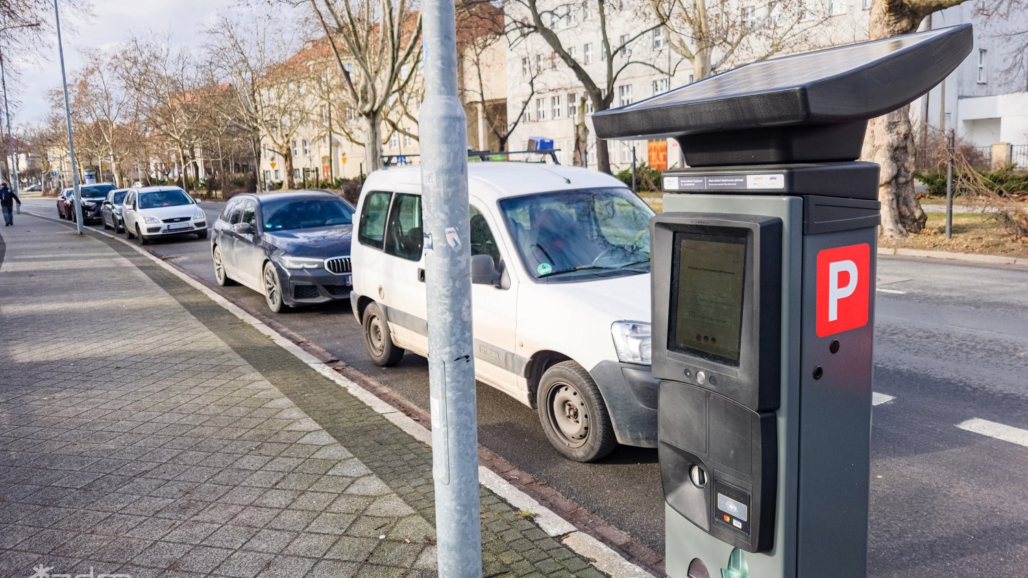 Szary parkomat stojący na chodniku. Za nim, w perspektywie, widać stojące samochody