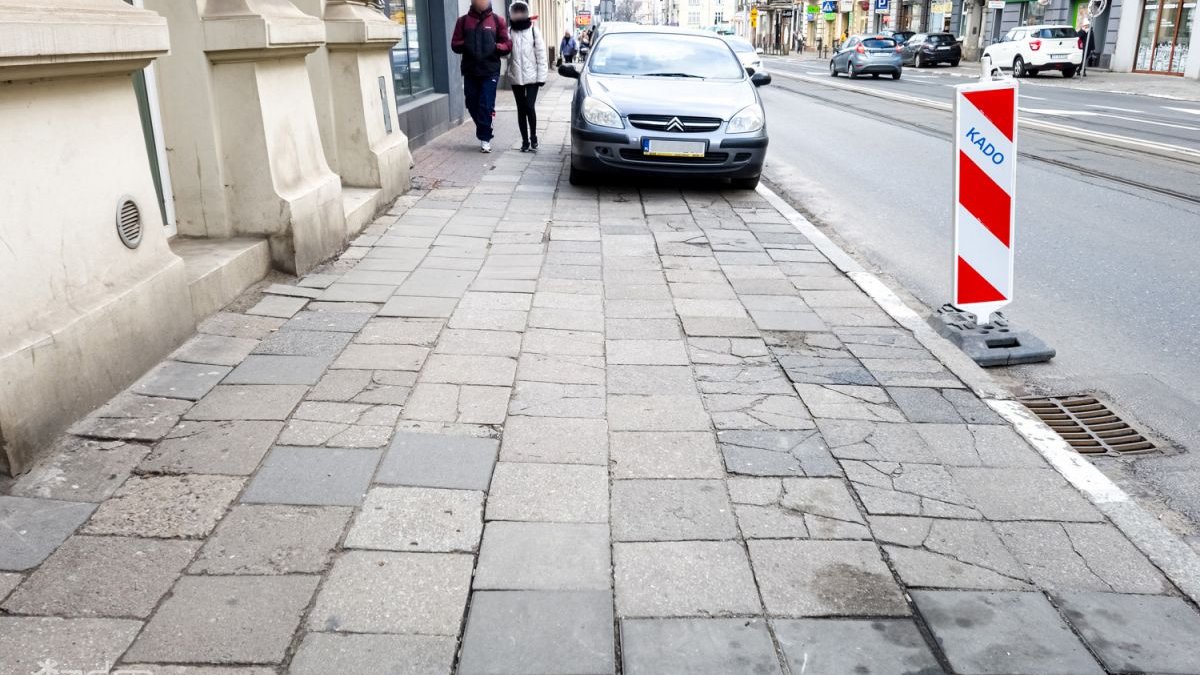 Chodnik ze zniszczonych płyteka przy ul. Dąbrowskiego przed remontem. Na chodniku zaparkowany samochód osóbowy, w wąskim przejściu obok auta idą dwie osoby.