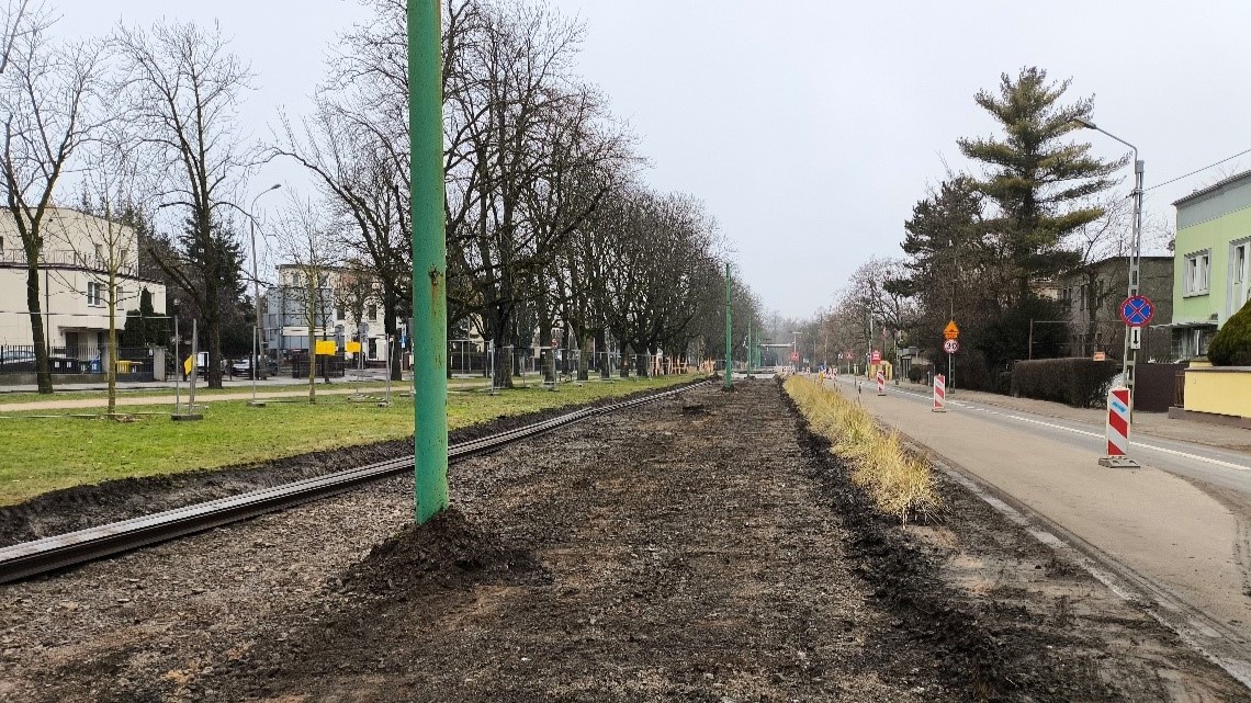 Miejsce po zdjętym torowisku wzdłuż al. Wielkopolskiej. Po prawej pas jezdni wygrodzony pylonami. Po lewej pas zieleni z drzewami. - grafika artykułu