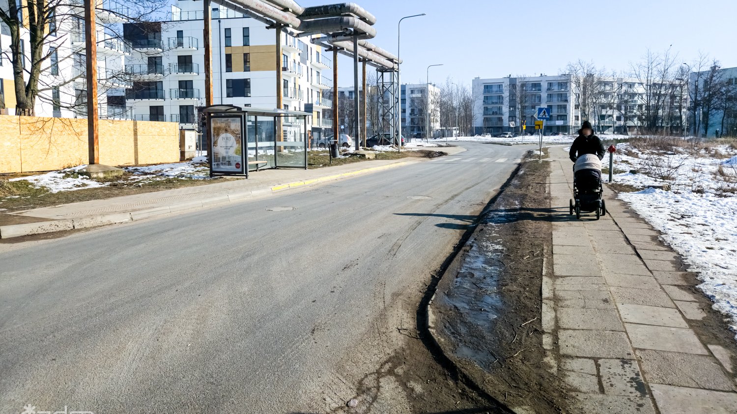 Zdjęcie z ul. Jasielskiej. W tle skrzyżowanie, które zmieni się w rondo. Po lewej przystanek autobusdowy, po prawej osóba z wózkiem dziecięcym na wąskim chodniku.