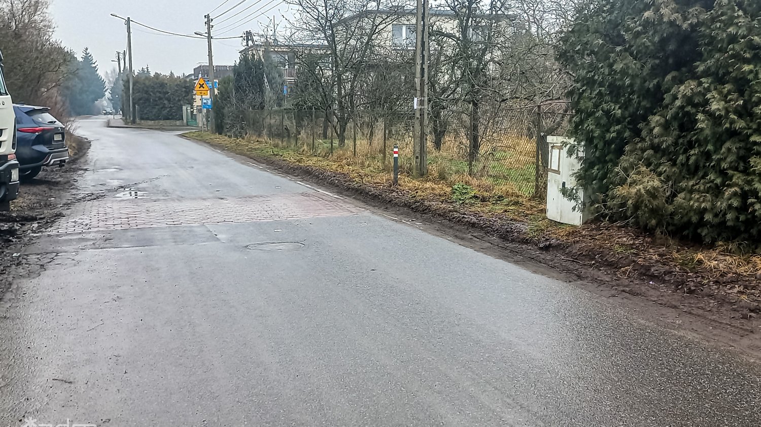 Zdjęcie ulic z Pokrzywna przed przebudową. Jezdnia asfaltowa bez chodnika, obok zieleń przyuliczna.