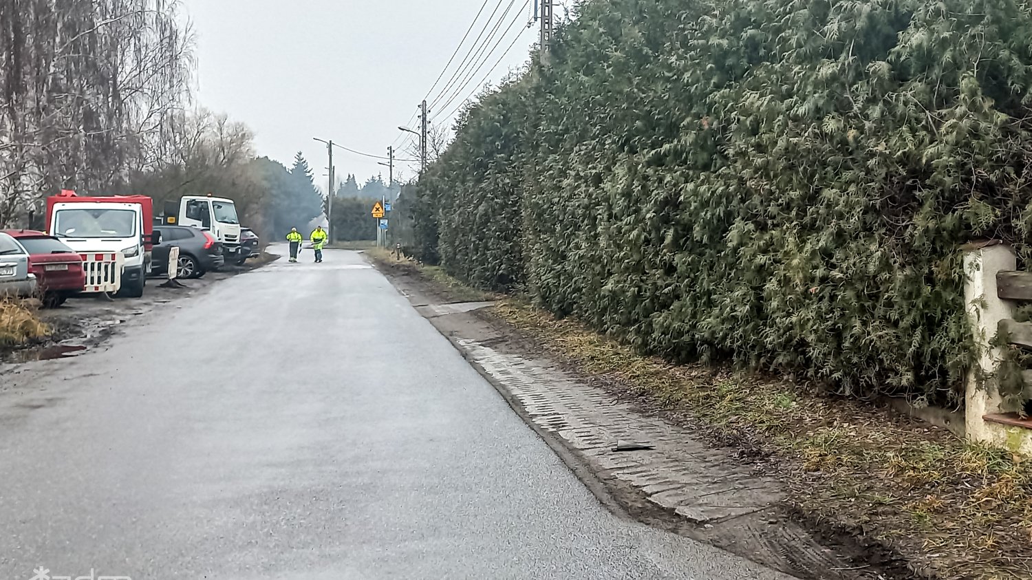 Zdjęcie ulic z Pokrzywna przed przebudową. Asfaltowa ulica bez chodnika, obok zieleń przyuliczna i parkujące pojazdy.