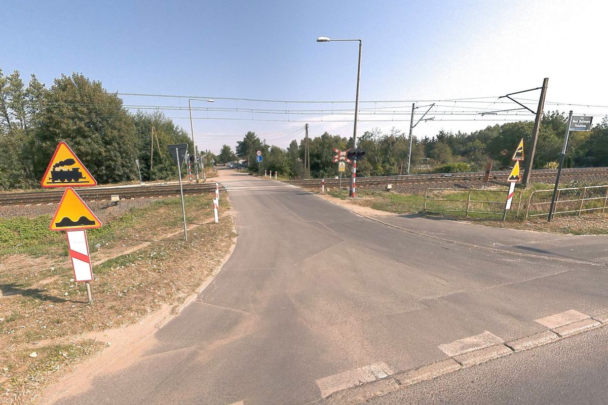 Zamknięcie przejazdu w ciągu ul. Nad Różanym Potokiem Tory kolejowe przecinają drogę asfaltową. Przy drodze stoją znaki ostrzegawcze, sygnalizacja świetlna oraz lampy uliczne. W tle widać drzewa, zarośla i kawałek nieba.